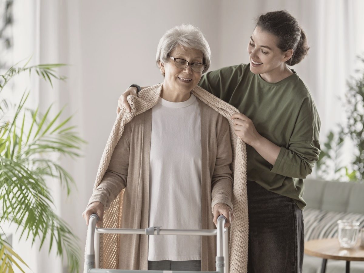 A caregiver helping an older woman with her sweater as she stands with her walker